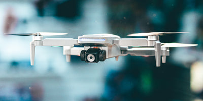 Sort Drone Recon XV1 i luften over en mark med grønne planter og træer i baggrunden.